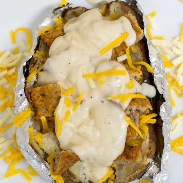Baked potato in foil, topped with white sauce, shredded cheese, and crispy chicken pieces.