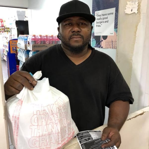 A man in a black cap and shirt holds a white takeout bag and a menu at a counter.