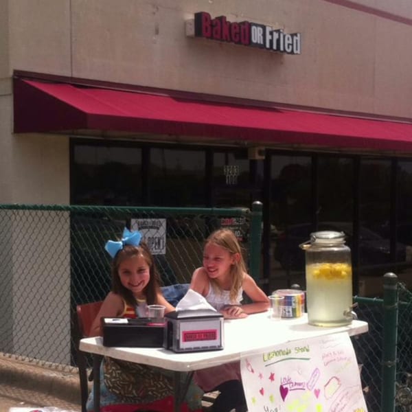 Two young girls smiling at a lemonade stand in front of the "Baked or Fried" restaurant.