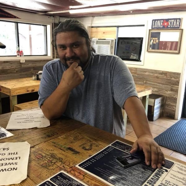 A man in a gray t-shirt smiles at the camera from behind a wooden counter with menus.