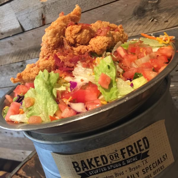 A large salad with fried chicken tenders, bacon bits, lettuce, and tomatoes in a metal bowl.