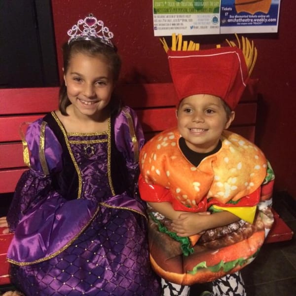 Two children in costumes, one a princess and the other a hamburger with fries, smiling on a red bench.