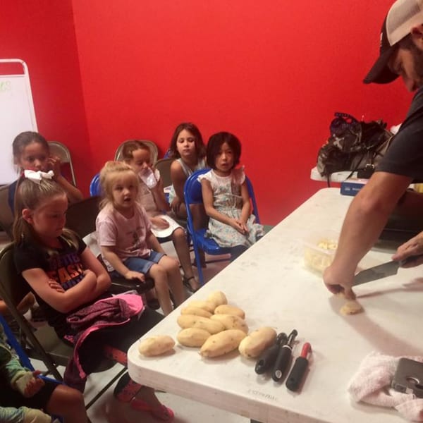 Children watch a chef prepare potatoes at a cooking class with a red wall background.